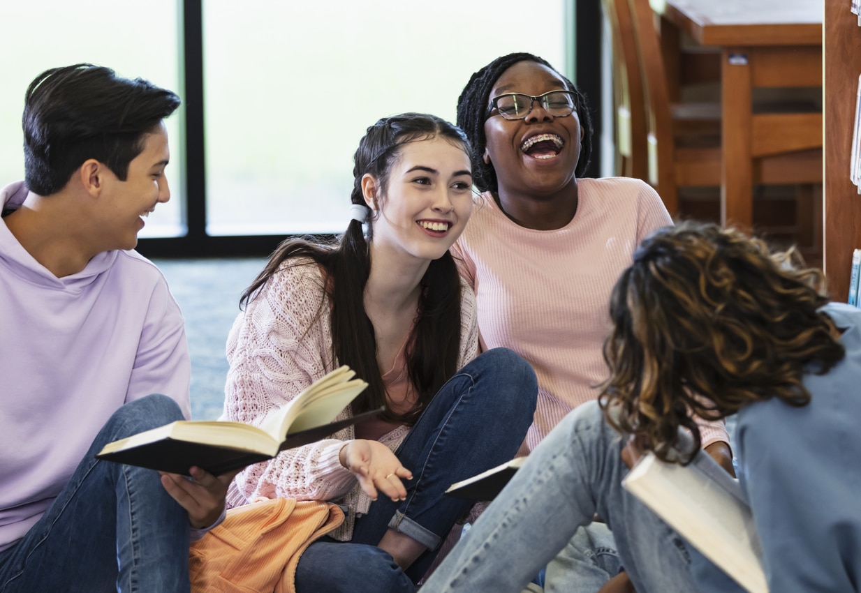 A multiracial group of four high school students studying together, sitting on the library floor with books. One girl is smiling as she speaks, and another girl is laughing.