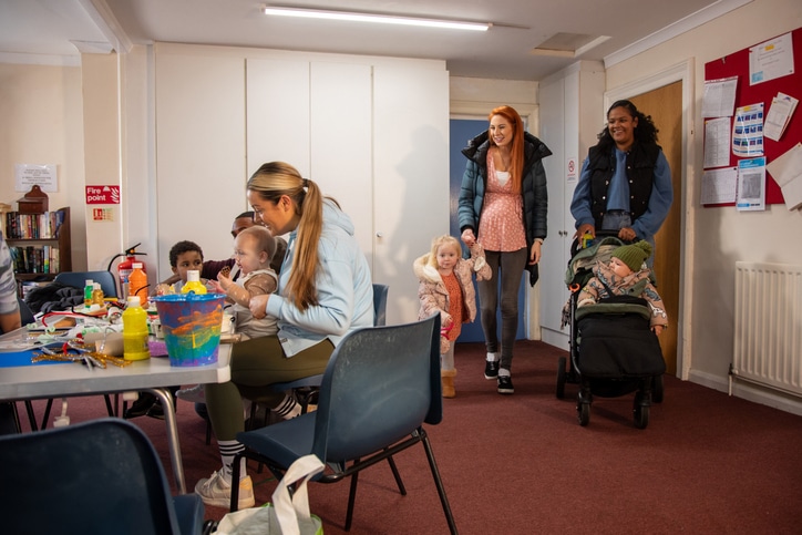 Full shot of two young female adults arriving at baby class with their children. There are adults and children already there. The institute is located in Seghill, Northumberland.