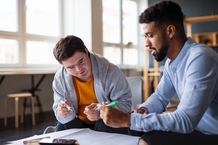 A young man with down syndrome is shown to be reviewing papers with a tutor in a classroom.